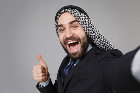 Close up of arabian muslim businessman in keffiyeh kafiya ring igal agal black suit isolated on gray background. Achievement career wealth business. Doing selfie shot on mobile phone showing thumb up.の写真素材