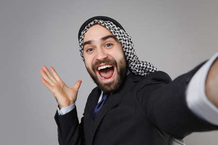 Close up of arabian muslim businessman in keffiyeh kafiya ring igal agal black suit isolated on gray background. Achievement career wealth business. Doing selfie shot on mobile phone spreading hands.の写真素材