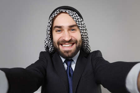 Close up of smiling arabian muslim businessman in keffiyeh kafiya ring igal agal black suit isolated on gray background. Achievement career wealth business concept. Doing selfie shot on mobile phone.の写真素材
