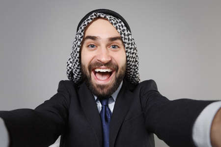 Close up of cheerful arabian muslim businessman in keffiyeh kafiya ring igal agal black suit isolated on gray background. Achievement career wealth business concept. Doing selfie shot on mobile phone.の写真素材