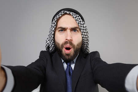 Close up of shocked arabian muslim businessman in keffiyeh kafiya ring igal agal black suit isolated on gray background. Achievement career wealth business concept. Doing selfie shot on mobile phone.の写真素材
