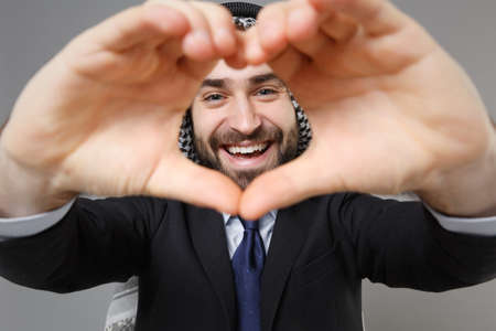 Smiling bearded arabian muslim businessman in keffiyeh kafiya ring igal agal classic black suit isolated on gray background. Achievement career wealth business concept. Showing shape heart with hands.の写真素材