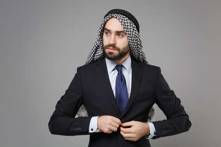 Handsome young bearded arabian muslim businessman in keffiyeh kafiya ring igal agal classic black suit shirt isolated on gray background. Achievement career wealth business concept. Fastening jacket.の写真素材