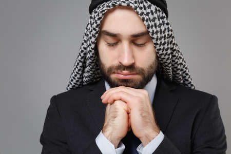 Close up of bearded arabian muslim businessman in keffiyeh kafiya ring igal agal classic suit isolated on gray background. Achievement career wealth business concept. Holding hands folded in prayer.の写真素材