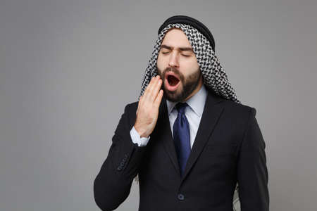 Tired young bearded arabian muslim businessman in keffiyeh kafiya ring igal agal classic suit isolated on gray background. Achievement career wealth business concept. Yawning covering mouth with hand.の写真素材