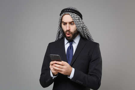 Shocked arabian muslim businessman in keffiyeh kafiya ring igal agal classic black suit isolated on gray background. Achievement career wealth business concept. Using mobile phone, typing sms message.の写真素材