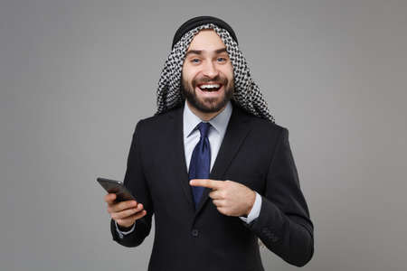 Funny arabian muslim businessman in keffiyeh kafiya ring igal agal classic black suit isolated on gray background. Achievement career wealth business concept. Pointing index finger on mobile phone.の写真素材