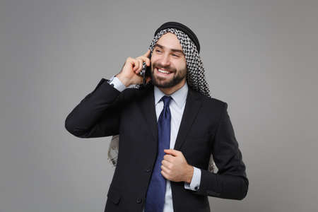 Smiling bearded arabian muslim businessman in keffiyeh kafiya ring igal agal classic black suit shirt isolated on gray background. Achievement career wealth business concept. Talking on mobile phone.の写真素材