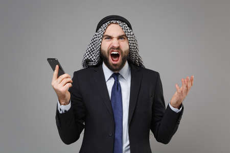 Irritated bearded arabian muslim businessman in keffiyeh kafiya ring igal agal classic black suit isolated on gray background. Achievement career wealth business concept. Talk on mobile phone scream.の写真素材