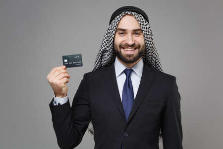Smiling bearded arabian muslim businessman in keffiyeh kafiya ring igal agal classic black suit shirt isolated on gray background. Achievement career wealth business concept. Hold credit bank card.の写真素材
