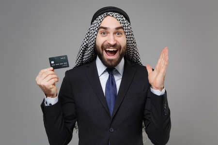 Excited bearded arabian muslim businessman in keffiyeh kafiya ring igal agal classic black suit shirt isolated on gray background. Achievement career wealth business concept. Hold credit bank card.の写真素材