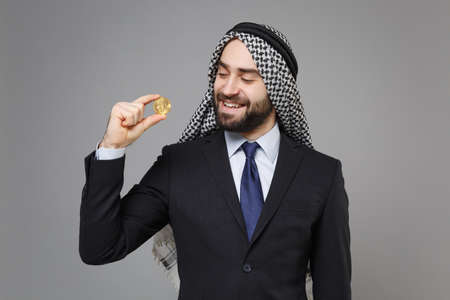 Smiling young bearded arabian muslim businessman in keffiyeh kafiya ring igal agal classic suit isolated on gray background. Achievement career wealth business concept. Hold bitcoin, future currency.の写真素材