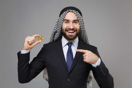 Funny arabian muslim businessman in keffiyeh kafiya ring igal agal classic suit isolated on gray background. Achievement career wealth business concept. Point index finger on bitcoin, future currency.の写真素材