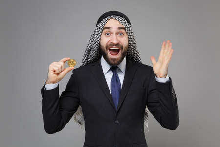 Surprised arabian muslim businessman in keffiyeh kafiya ring igal agal black suit isolated on gray background. Achievement career wealth business concept. Hold bitcoin future currency spreading hands.の写真素材