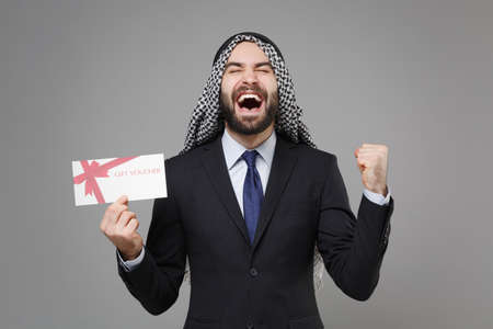 Overjoyed bearded arabian muslim businessman in keffiyeh kafiya ring igal agal suit isolated on gray background. Achievement career wealth business concept. Hold gift certificate doing winner gesture.の写真素材