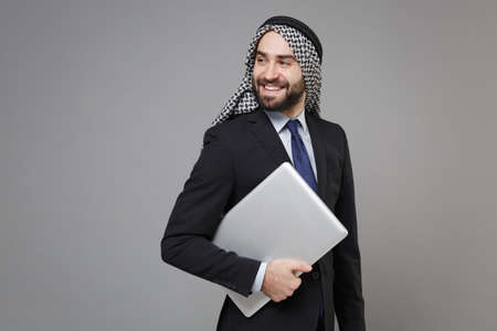 Successful young bearded arabian muslim businessman in keffiyeh kafiya ring igal agal classic black suit isolated on gray background. Achievement career wealth business concept. Hold laptop computer.の写真素材