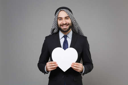Smiling bearded arabian muslim businessman in keffiyeh kafiya ring igal agal classic suit isolated on gray background. Achievement career wealth business concept. Hold white heart with copy space.の写真素材