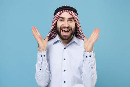 Excited bearded young arabian muslim man in keffiyeh kafiya ring igal agal casual clothes isolated on pastel blue background. People religious lifestyle concept. Mock up copy space. Spreading hands.の写真素材