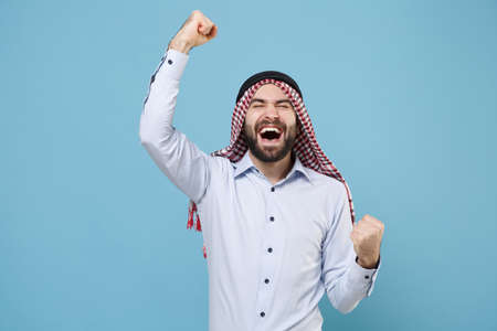 Happy young arabian muslim man in keffiyeh kafiya ring igal agal casual clothes isolated on pastel blue background. People religious lifestyle concept. Mock up cope space. Clenching fists like winner.の写真素材