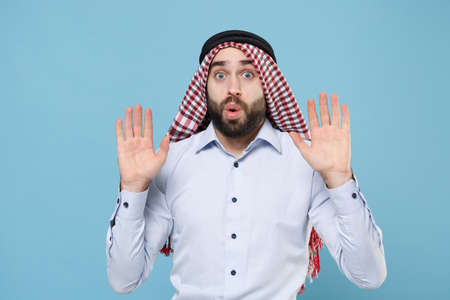 Shocked young bearded arabian muslim man in keffiyeh kafiya ring igal agal casual clothes posing isolated on pastel blue background. People religious lifestyle concept. Rising hands up, showing palms.の写真素材