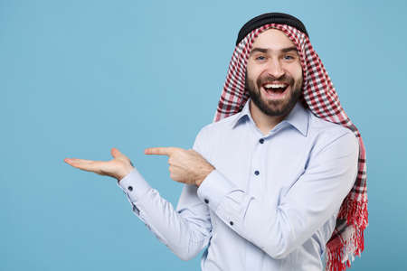 Cheerful young bearded arabian muslim man in keffiyeh kafiya ring igal agal casual clothes isolated on pastel blue background. People religious lifestyle concept. Pointing index finger hand aside.の写真素材
