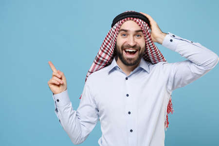 Amazed bearded arabian muslim man in keffiyeh kafiya ring igal agal casual clothes isolated on pastel blue background. People religious lifestyle concept. Point index finger aside up put hand on head.の写真素材