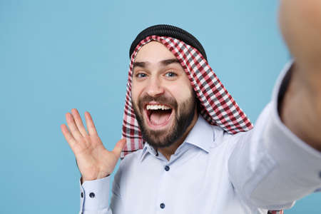 Close up of funny bearded arabian muslim man in keffiyeh kafiya ring igal agal casual clothes isolated on pastel blue background. People religious lifestyle concept. Doing selfie shot on mobile phone.の写真素材