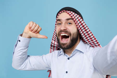 Close up of arabian muslim man in keffiyeh kafiya ring igal agal casual clothes isolated on pastel blue background. People religious concept. Doing selfie shot on mobile phone, point thumb on himself.の写真素材