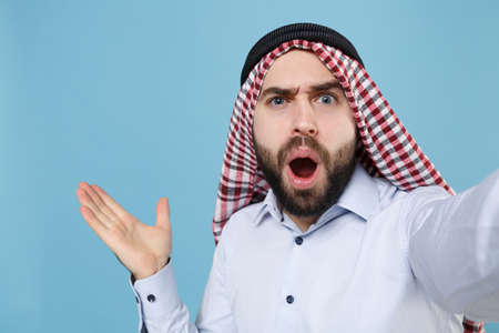 Close up of shocked young arabian muslim man in keffiyeh kafiya ring igal agal casual clothes isolated on pastel blue background. People religious lifestyle concept. Doing selfie shot on mobile phone.の写真素材