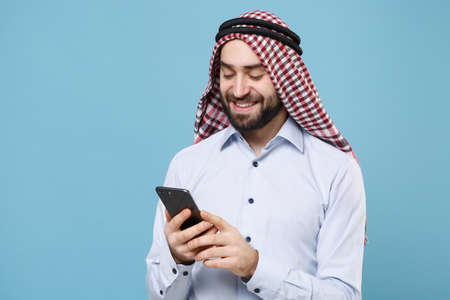Smiling young bearded arabian muslim man in keffiyeh kafiya ring igal agal casual clothes isolated on pastel blue background. People religious lifestyle concept. Using mobile phone typing sms message.の写真素材