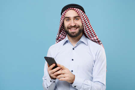 Smiling young bearded arabian muslim man in keffiyeh kafiya ring igal agal casual clothes isolated on pastel blue background. People religious lifestyle concept. Using mobile phone typing sms message.の写真素材