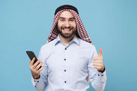 Smiling young bearded arabian muslim man in keffiyeh kafiya ring igal agal casual clothes isolated on pastel blue background. People religious lifestyle concept. Hold mobile phone, showing thumb up.の写真素材