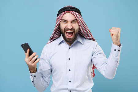Happy young bearded arabian muslim man in keffiyeh kafiya ring igal agal casual clothes isolated on pastel blue background. People religious lifestyle concept. Hold mobile phone, doing winner gesture.の写真素材