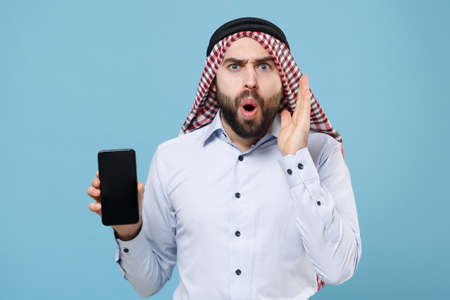 Shocked bearded arabian muslim man in keffiyeh kafiya ring igal agal casual clothes isolated on pastel blue background. People religious lifestyle concept. Hold mobile phone with blank empty screen.の写真素材