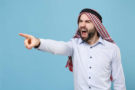 Angry irritated arabian muslim man in keffiyeh kafiya ring igal agal casual clothes isolated on pastel blue background. People religious lifestyle concept. Point index finger aside swearing screaming.の写真素材
