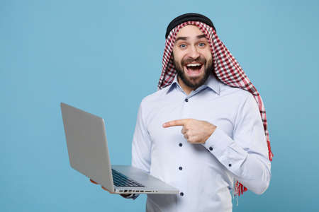 Excited bearded arabian muslim man in keffiyeh kafiya ring igal agal casual clothes isolated on pastel blue background. People religious lifestyle concept. Pointing index finger on laptop pc computer.の写真素材
