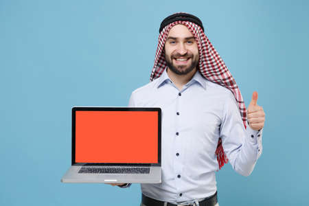 Smiling arabian muslim man in keffiyeh kafiya ring igal agal casual clothes isolated on pastel blue background. People religious concept. Hold laptop computer with blank empty screen showing thumb up.の写真素材