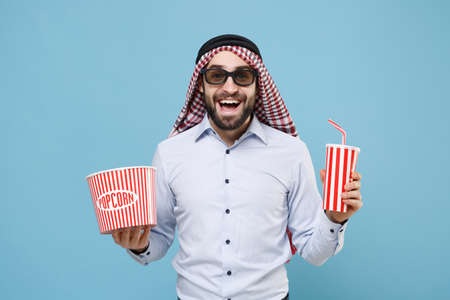 Cheerful arabian muslim man in keffiyeh kafiya ring igal agal 3d  glasses isolated on pastel blue background. People religious concept. Watching movie film, holding bucket of popcorn, cup of soda.の写真素材