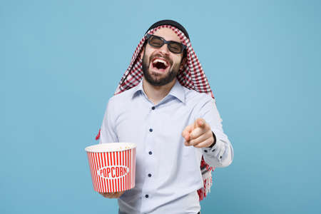 Laughing arabian muslim man in keffiyeh kafiya ring igal agal 3d  glasses isolated on pastel blue background. People religious concept. Watching movie film hold bucket of popcorn point on camera.の写真素材