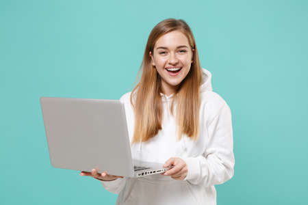 Cheerful young woman girl in casual white hoodie posing isolated on blue turquoise background studio portrait. People emotions lifestyle concept. Mock up copy space. Working on laptop pc computer.の写真素材