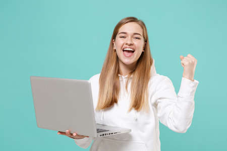 Happy young woman girl in casual white hoodie posing isolated on blue turquoise background studio portrait. People lifestyle concept. Mock up copy space. Hold laptop pc computer, doing winner gesture.の写真素材