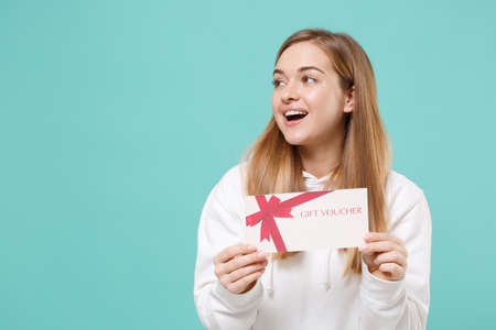 Funny young woman girl in casual white hoodie posing isolated on blue turquoise wall background studio portrait. People lifestyle concept. Mock up copy space. Hold gift certificate, looking aside.の写真素材
