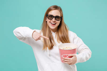 Funny young woman in casual white hoodie 3d glasses isolated on blue turquoise background. People lifestyle concept. Mock up copy space. Watching movie film pointing index finger on bucket of popcorn.の写真素材
