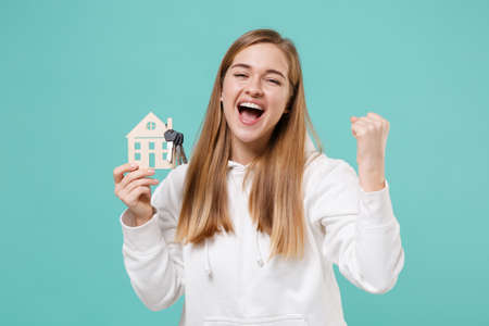 Happy young woman girl in casual white hoodie posing isolated on blue turquoise background studio portrait. People lifestyle concept. Mock up copy space. Hold house bunch of keys doing winner gesture.の写真素材