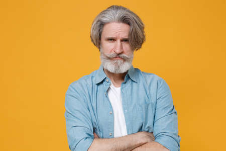Dissatisfied elderly gray-haired mustache bearded man in casual blue shirt posing isolated on yellow...