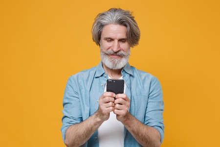 Smiling elderly gray-haired mustache bearded man in casual blue shirt posing isolated on yellow background studio.の写真素材