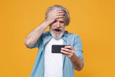 Shocked elderly gray-haired mustache bearded man in casual blue shirt posing isolated on yellow background.の写真素材
