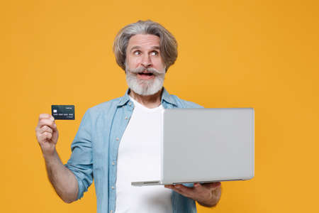 Pensive elderly gray-haired mustache bearded man in casual blue shirt posing isolated on yellow wall...