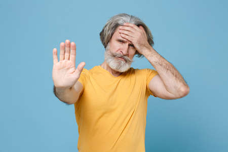 Tired elderly gray-haired mustache bearded man in casual yellow t-shirt isolated on blue wall background.の写真素材