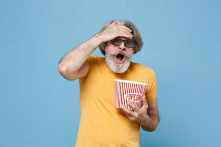 Scared elderly gray-haired mustache bearded man in yellow t-shirt 3d glasses isolated on blue background. People emotions in cinema concept. Watching movie film hold bucket popcorn put hand on head.の写真素材